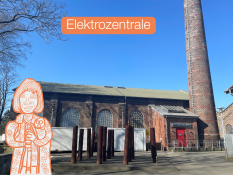 Auf dem Bild sieht man ein großes Backstein‑Gebäude. Vor dem Gebäude stehen viele hohe, dunkle Metallpfosten. Rechts neben dem Gebäude ist ein hoher Schornstein aus Backstein. Am Himmel ist keine Wolke zu sehen, es ist klar und blau. Auf dem Bild steht das Wort „Elektrozentrale“ in einem orangefarbenen Kasten. Links im Bild ist eine gezeichnete Figur zu sehen, die einen Blindenstock in den Händen hält.