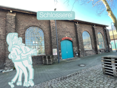 Bildbeschreibung:
Auf dem Bild ist ein langes Gebäude aus dunklen Backsteinen zu sehen. Es hat drei große, bogenförmige Fenster. In der Mitte befindet sich eine große, blau gestrichene Tür. Über der Tür hängt ein rotes Schild mit dem Wort „Schlosserei“. Links im Bild stehen zwei miteinander tanzende Figuren. Vor dem Gebäude liegen Holzpaletten. Der Boden besteht aus Kopfsteinpflaster. Der Himmel ist blau und klar.