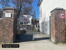 Auf dem Bild sieht man einen Eingang zwischen zwei alten Backstein‑Pfeilern. In der Mitte steht ein Metalltor, das geöffnet ist. Am Tor hängt ein großes Banner mit der Aufschrift „Zentrum Altenberg“. Links und rechts stehen Schilder an den Mauern. Hinter dem Tor führt ein Weg in eine Grünanlage. Der Himmel ist blau und die Sonne scheint. Unten im Bild steht der Text „Eingang vorne“ in einem schwarzen Kasten.