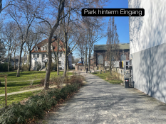Auf dem Bild sieht man einen breiten Spazierweg. Er führt durch einen kleinen Park mit Grasflächen und Bäumen. Links stehen alte Gebäude mit großen Fenstern und einem roten Dach.  Oben im Bild steht der Text „Park hinterm Eingang“ in einem schwarzen Kasten.