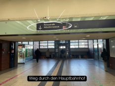 Auf dem Bild sieht man einen Ausgang aus dem Hauptbahnhof. Der Weg führt nach draußen auf eine offene Fläche. Rechts stehen mehrere schräg eingebaute Metallstangen neben einer Backsteinmauer mit Graffiti. Links liegt eine Grünfläche mit Gras und einem kleinen Hügel. Auf dem Boden verläuft eine grüne Linie, die den Weg markiert. Unten im Bild steht der Text „Ausgang Hauptbahnhof“ in einem schwarzen Kasten.