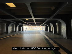 Auf dem Bild sieht man einen Tunnel im Hauptbahnhof. Der Bereich ist dunkel, aber am Ende des Tunnels scheint helles Tageslicht. Dort ist der Ausgang zu sehen. Der Boden hat helle und dunkle Fliesen sowie Linien auf dem Boden. Im Hintergrund gehen einige Personen in Richtung Ausgang. Unten im Bild steht der Text „Weg durch den HBF Richtung Ausgang“ in einem schwarzen Kasten.