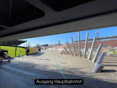 Auf dem Bild sieht man einen Ausgang aus dem Hauptbahnhof. Der Weg führt nach draußen auf eine offene Fläche. Rechts stehen mehrere schräg eingebaute Metallstangen neben einer Backsteinmauer mit Graffiti. Links liegt eine Grünfläche mit Gras und einem kleinen Hügel. Auf dem Boden verläuft eine grüne Linie, die den Weg markiert. Unten im Bild steht der Text „Ausgang Hauptbahnhof“ in einem schwarzen Kasten.