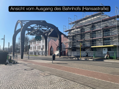 Auf dem Bild sieht man eine Straße direkt am Ausgang des Bahnhofs. Links steht eine große Metallkonstruktion mit Bögen. Dahinter befindet sich ein weißes Gebäude mit dem Schriftzug „ALTENBERG“. Rechts steht ein großes Gebäude, das gerade eingerüstet ist. Der Himmel ist klar und blau. Unten im Bild steht der Text „Ansicht vom Ausgang des Bahnhofs (Hansastraße)“ in einem schwarzen Kasten.