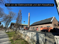 Auf dem Bild sieht man einen Teil eines Parks mit Bäumen und Wegen. Dahinter stehen zwei große Backstein‑Gebäude. Eines hat einen hohen Schornstein. Vor den Gebäuden stehen Autos und Lampen. Der Himmel ist klar und blau. Oben im Bild steht der Text „Park vor der Elektrozentrale und dem Kesselhaus“ in einem schwarzen Kasten.