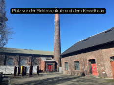 Auf dem Bild sieht man einen offenen Platz zwischen zwei Backstein‑Gebäuden. In der Mitte steht ein sehr hoher Schornstein aus roten Ziegeln. Beide Gebäude haben rote Türen und große Fenster. Der Himmel ist klar und blau. Auf dem Bild steht oben der Text „Platz vor der Elektrozentrale und dem Kesselhaus“ in einem schwarzen Kasten.