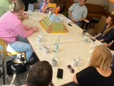 Beteiligung: Mehrere Personen sitzen an einem Tisch, auf dem viele Tassen und Gläser stehen. Im Hintergrund steht ein großer Fernseher und in der Tischmitte die Partizipationstreppe. Bei dieser Gruppe handelt es sich um die Gruppe der Echologen.