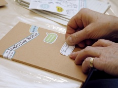 Methoden: Eine Person klebt Sticker mit der Aufschrift Lass mal gemeinsam machen auf ein braunes Notizbuch mit der Beschriftung Inklusive Kultur.