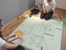 Methoden: Im Mittelpunkt ist ein großes Plakat zu sehen, welches auf einem Holzboden ausgebreitet ist. Drum herum sitzen Maximiliane Horbach und Hedda Langenberg und zeichnen mehrere Türen auf das Plakat. Im Hintergrund befindet sich ein Stehtisch und ein Fenster.