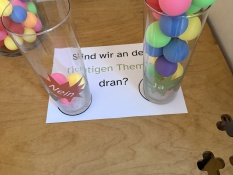 Methoden: Auf einem Holztisch stehen zwei transparente Behälter mit den Beschriftungen  ja und nein. In dem Ja-Behälter befinden sich viele bunte Kugeln, während der Nein-Behälter leer ist. Darunter liegt ein Blatt Papier mit der Frage Sind wir an den richtigen Themen dran?