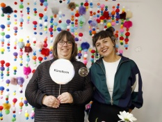 Zwei Personen stehen lächelnd vor einer Wand voller bunter Pompons. Die Person links hält ein rundes Schild mit der Aufschrift Künstlerin in den Händen. Vor ihnen schweben Seifenblasen.
