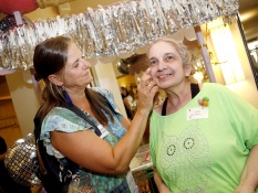 Zwei Frauen stehen nebeneinander. Die linke Frau schminkt die rechte Frau im Gesicht mit Glitzer. Beide lächeln und im Hintergrund sieht man silberne Girlanden.