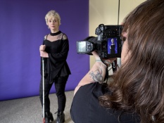 Im Mittelpunkt des Fotos steht Nadine Rockstein, die in eine Kamera schaut. Sie hält einen Langstock in der Hand und posiert für die Kamera. Im Hintergrund sieht man einen blauen Hintergrund und hinter der Kamera steht die Fotografin Anna Spindelndreier.