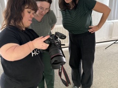 Drei Frauen stehen nebeneinander und betrachten gemeinsam das Display einer Kamera. Diese Kamera hält die Fotografin Anna Spindelndreier in der Hand. Im Hintergrund sind graue Vorhänge zu sehen und ein grauer Boden.