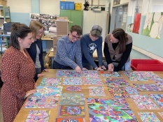 In einer Werkstatt für Menschen mit Behinderung betrachten 5 Personen gemalte Bilder, die vor ihnen auf dem Tisch liegen. Die Bilder sind bunt gestaltet mit vielen detaillierten geometrischen Figuren.