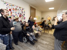 Vor einer Wand aus bunten Puscheln steht eine Gruppe von 5 Personen im Projektbüro. Einige von ihnen halten bunte Schilder hoch, auf denen Sprüche wie Alle für Alle und The future is inclusive (Die Zukunft ist inklusiv) abgebildet sind. Rechts im Bild befindet sich eine Frau, die ein Foto von der Gruppe macht. Im Hintergrund sitzen drei weitere Personen auf einem Sofa und unterhalten sich.