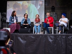 Eine Gruppe von 5 Personen sitzt in einer Reihe auf der OpernAir-Bühne beim Bürger*innenfest des Caritasverbandes Oberhausen Eine Party für Alle und unterhalten sich. Von links nach rechts sitzen dort: Bianca Mehanovic, Barbara Klingels, Jennifer Lorenz, Claudia Kosch und Denise Maurischat. Im Hintergrund ist auf einer Leinwand ein Bild von dem Workshop Demokratiesäulen bauen zu sehen und drei Personen, die mit Holz arbeiten.