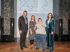 Vier Personen stehen in eleganter Kleidung nebeneinander. Im Hintergrund steht auf einer Leinwand Kulturgestalten. Alle lächeln in die Kamera und Jochen Riemer hält den Zukunftspreis KULTURGESTALTEN 2024 der kulturpolitischen Gesellschaft in der Hand.