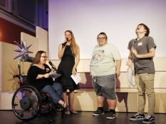 Vier Personen stehen auf einer Bühne und lächeln. Eine von ihnen hält ein Mikrofon in der Hand und redet zum Publikum. Ganz rechts sitzt eine Frau im Rollstuhl