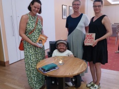 Romi Domkowsky, Bianca Mehanovic und Claudia Kosch stehen gemeinsam um einen Tisch herum. In ihrer Mitte sitzt der Aktivist Raul Krauthausen in einem elektrischen Rollstuhl. Bianca hält sein Buch mit dem Titel Wer Inklusion will, findet einen Weg. Wer sie nicht will, findet Ausreden in den Händen.