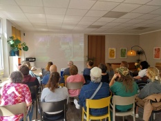 Eine Gruppe von Menschen sitzt im Projektbüro auf Stühlen in mehreren Reihen in einer Kinosituation. Sie betrachten alle eine Projektion an der Wand. Auf diesem projizierten Bild sieht man eine Frau, die einer Person im Rollstuhl eine Tür aufhält.
