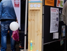 Auf dem Bild sieht man eine selbstgebaute Demokratiesäule vor einem Stand des Caritasverbandes Oberhausen beim Bürger*innenfest. Bei dieser Säule handelt es sich um eine Holzkonstruktion, die aus drei durchsichtigen Röhren besteht. Darunter steht eine Kiste mit bunten Tischtennisbällen, die man in die Röhren werfen kann. Oben drüber auf einem Zettel befindet sich die Aussage: Oberhausen ist die schönste Stadt weit und breit. Eine Antwort auf diese Frage kann man geben, indem man einen Ball entweder in die Röhre mit einem Daumen hoch, einem Daumen runter oder einem mittleren Daumen wirft.