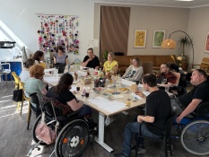 Eine Gruppe von 11 Leuten sitzt an einem großen Tisch im Projektbüro. Auf dem Tisch vor ihnen befinden sich mehrere Gläser mit Getränken und Snacks wie zum Beispiel Salzstangen.