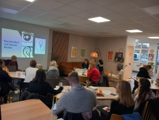Im Projektbüro sitzen mehrere Menschen an Tischen. Sie hören Nadine Rockstein zu, die vorne an einem Laptop sitzend etwas erklärt. Auf der Wand hinter ihr wird eine Präsentation gezeigt. Darauf steht: Barrierefrei auf Social Media. Man sieht Bilder von einem Auge, einem Ohr und einer Glühbirne.