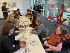 n einem langen Tisch sitzen Teilnehmer*innen einer Arbeitsgruppe. Sie haben Schilder mit Zeichnungen vor sich liegen. Im Hintergrund steht der Kameramann Jan Krämer und filmt mit. Das gesamte Treffen findet in der Ludwiggalerie im Schloss Oberhausen statt.