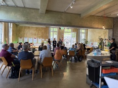 In einem hellen Raum sitzen mehrere Personen an Tischen. Sie hören einer kleinen Gruppe von Vortragenden vorne im Raum zu. Im Raum befinden sich viele Fenster und im Hintergrund sind farbige Poster zu sehen. Rechts befindet sich eine Theke.